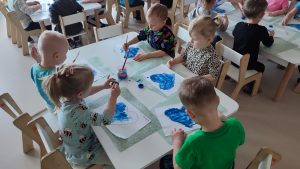 Dzieci siedzą przy białych stolikach i malują serduszko niebieską farbą.