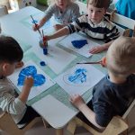 Dzieci siedzą przy białych stolikach i malują serduszko niebieską farbą.