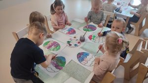 Dzieci siedzą przy białych stolikach, na których rozłożone są zielone podkładki. Na nich kontur pisanki, który dzieci malują farbami.