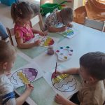 Dzieci siedzą przy białych stolikach, na których rozłożone są zielone podkładki. Na nich kontur pisanki, który dzieci malują farbami.