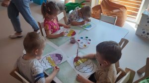 Dzieci siedzą przy białych stolikach, na których rozłożone są zielone podkładki. Na nich kontur pisanki, który dzieci malują farbami.