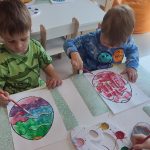 Dzieci siedzą przy białych stolikach, na których rozłożone są zielone podkładki. Na nich kontur pisanki, który dzieci malują farbami.