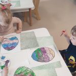 Dzieci siedzą przy białych stolikach, na których rozłożone są zielone podkładki. Na nich kontur pisanki, który dzieci malują farbami.