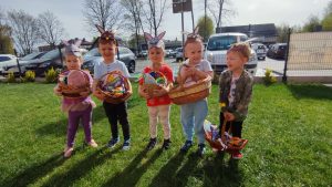 5 dzieci stoi na zielonej trawie, w rękach trzymają wielkanocne koszyczki. Na głowach mają czapki zajączka.