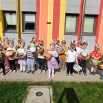 Duża grupa dzieci pozuje do zdjęcia na tle budynku w kolorowe paski. Dzieci w rękach trzymają wielkanocne koszyczki.