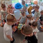 grupa dzieci na szarym dywanie stoją wokół dużego pudła, w którym znajdują się kwadratowe prezenty.
