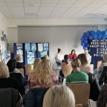 Rodzice i nauczyciele siedzący w sali przedszkolnej. Naprzeciwko, za stołem siedzą trzy Panie prowadzące warsztaty. W tle, po lewej stronie widać prace plastyczne, po prawej stronie znajduje się tablica z napisami oraz niebieskimi balonami.