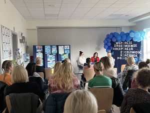 Rodzice i nauczyciele siedzący w sali przedszkolnej. Naprzeciwko, za stołem siedzą trzy Panie prowadzące warsztaty. W tle, po lewej stronie widać prace plastyczne, po prawej stronie znajduje się tablica z napisami oraz niebieskimi balonami.