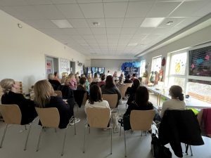 Rodzice i nauczyciele siedzący w sali przedszkolnej. Naprzeciwko, za stołem, siedzą trzy Panie prowadzące warsztaty. W tle, po lewej stronie widać prace plastyczne, po prawej stronie znajduje się tablica z napisami oraz niebieskimi balonami.