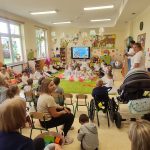 Dzieci siedzą na krzesełkach i patrzą na występy przedszkolaków z Przedszkola nr 19.