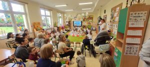Dzieci siedzą na krzesełkach i patrzą na występy przedszkolaków z Przedszkola nr 19.