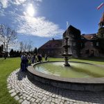 Grupa dzieci z opiekunami stoi przy fontannie na tle zamku w Gutowie.
