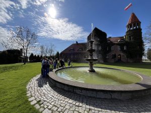 Grupa dzieci z opiekunami stoi przy fontannie na tle zamku w Gutowie.