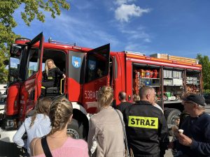 Dziewięć dorosłych osób stojących naprzeciwko wozu strażackiego, w którym siedzi Pani Strażak