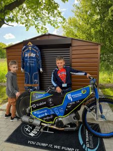 Dwóch chłopców stojących koło motocykla żużlowego. W tle wisi strój żużlowca.