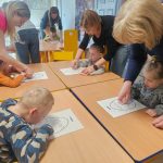 Dzieci siedzą przy stolikach i kreślą znaki na muzogramie zgodnie z muzyką.