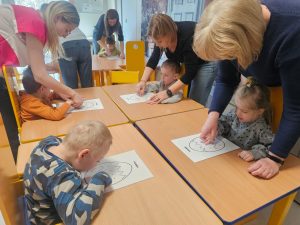 Dzieci siedzą przy stolikach i kreślą znaki na muzogramie zgodnie z muzyką.