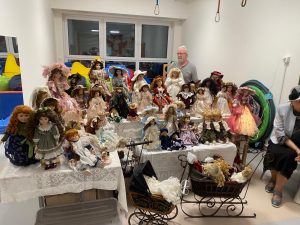 Kolekcja porcelanowych lalek ułożone na białych stołach. Obok stoi starszy mężczyzna.
