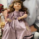 Starsza kobieta w szarej bluzce trzyma w rękach lalkę z porcelany ubraną w fioletową sukienkę.