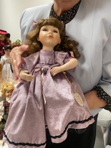Starsza kobieta w szarej bluzce trzyma w rękach lalkę z porcelany ubraną w fioletową sukienkę.