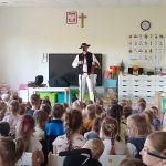 Grupa dzieci siedzi i patrzy na górala, który prezentuje strój góralski.