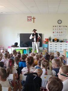 Grupa dzieci siedzi i patrzy na górala, który prezentuje strój góralski.