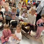 Grupa dzieci pozuje do zdjęcia z lalkami z porcelany . Obok stoi starsza kobieta w siwych włosach.
