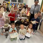 Grupa dzieci pozuje do zdjęcia z lalkami z porcelany . Obok stoi starsza kobieta w siwych włosach.