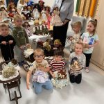 Grupa dzieci pozuje do zdjęcia z lalkami z porcelany . Obok stoi starsza kobieta w siwych włosach.