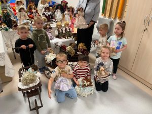 Grupa dzieci pozuje do zdjęcia z lalkami z porcelany . Obok stoi starsza kobieta w siwych włosach.