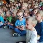 Duża grupa dzieci siedzi na szarym dywanie i uczestniczy w koncercie muzyki góralskiej.