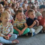Duża grupa dzieci siedzi na szarym dywanie i uczestniczy w koncercie muzyki góralskiej.