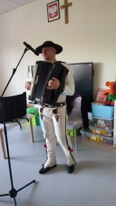 Mężczyzna ubrany w strój góralski gra na akordeonie. Stoi obok białego stolika.