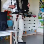 Mężczyzna ubrany w strój góralski gra na akordeonie. Stoi obok białego stolika.