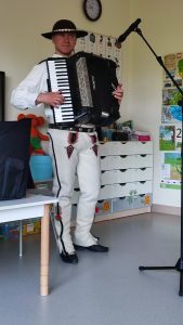 Mężczyzna ubrany w strój góralski gra na akordeonie. Stoi obok białego stolika.