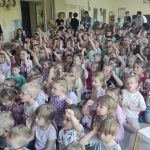 Grupy dzieci siedzące na dywanie w sali przedszkolnej. Część dzieci trzyma rękę podniesioną do góry. Z tyłu i z boku stoją panie.