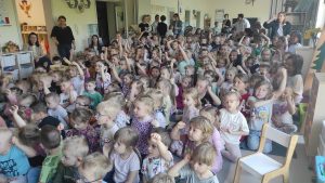 Grupy dzieci siedzące na dywanie w sali przedszkolnej. Część dzieci trzyma rękę podniesioną do góry. Z tyłu i z boku stoją panie.