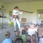 Góral stojący na środku sali, trzymający instrument muzyczny. Naprzeciwko muzyka siedzą dzieci.