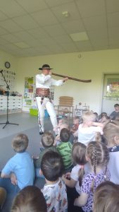 Góral stojący na środku sali, trzymający instrument muzyczny. Naprzeciwko muzyka siedzą dzieci.