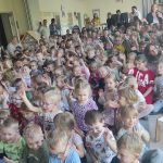 Grupy dzieci siedzące na dywanie w sali przedszkolnej. Część dzieci trzyma rękę podniesioną do góry. Z tyłu i z boku stoją panie.