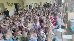 Grupy dzieci siedzące na dywanie w sali przedszkolnej. Część dzieci trzyma rękę podniesioną do góry. Z tyłu i z boku stoją panie.