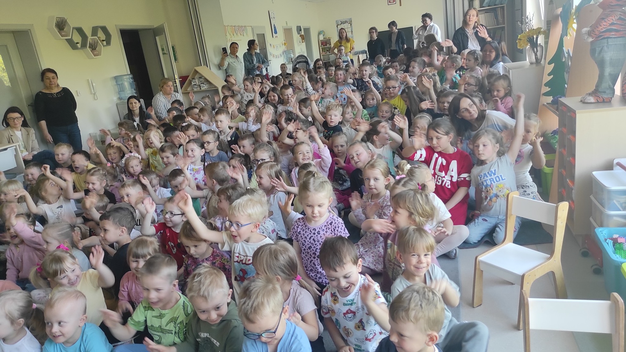 Grupy dzieci siedzące na dywanie w sali przedszkolnej. Część dzieci trzyma rękę podniesioną do góry. Z tyłu i z boku stoją panie.