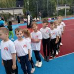 Dziewięcioro dzieci stojących w jednym rzędzie na boisku sportowym. Dzieci mają ubrane koszulki z logo przedszkola. W tle biegające dzieci i osoby dorosłe.