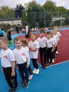 Dziewięcioro dzieci stojących w jednym rzędzie na boisku sportowym. Dzieci mają ubrane koszulki z logo przedszkola. W tle biegające dzieci i osoby dorosłe.