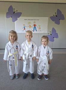 Troje dzieci ubranych w stroje do Teakwondo, stojących w sali przedszkolnej.