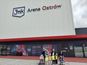 Sześcioro dzieci i cztery Panie pozujące do zdjęcia przed budynkiem 3mk Arena Ostrów. Dwoje dzieci siedzi w wózkach.