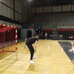Dziewczynka, wraz z Panią stojący naprzeciwko bramki na hali sportowej. W bramce stoi sportowiec. W tle stoją trzy osoby dorosłe.