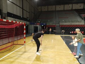 Dziewczynka, wraz z Panią stojący naprzeciwko bramki na hali sportowej. W bramce stoi sportowiec. W tle stoją trzy osoby dorosłe.