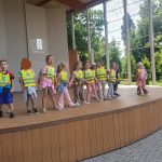 Grupa dzieci stoi na estradzie w parku, śpiewają piosenki.