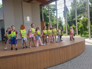 Grupa dzieci stoi na estradzie w parku, śpiewają piosenki.